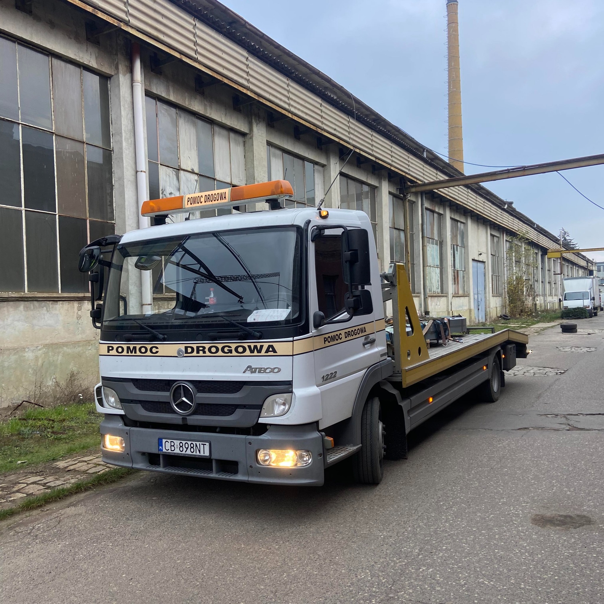 Biała laweta Mercedes Atego z napisem 'Pomoc Drogowa' na tle starej hali przemysłowej z ceglanym kominem w tle.