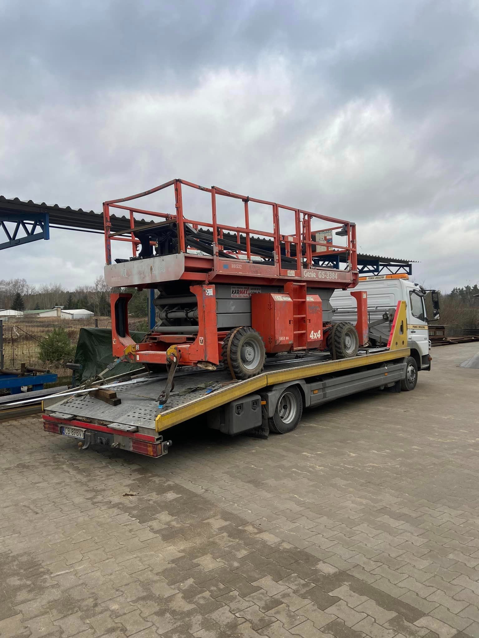 Pomarańczowy podnośnik nożycowy Genie GS-3384, z oznaczeniem 4x4, zabezpieczony pasami, przewożony na platformie lawety z żółtymi elementami, na tle pochmurnego nieba i przemysłowego otoczenia.