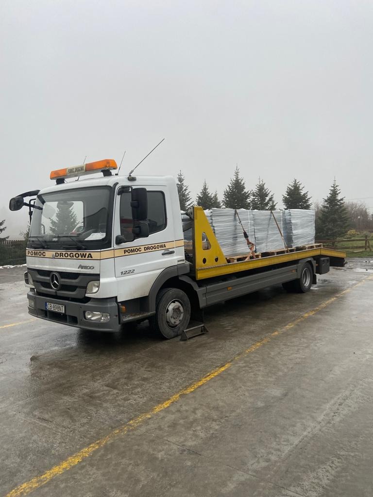 Ciężarówka pomocy drogowej Mercedes Atego 1222 z załadowanymi paletami na żółtej platformie, zaparkowana na mokrym placu z widocznymi drzewami w tle.