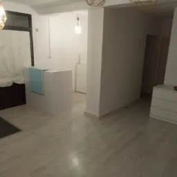 Jasne, minimalistyczne wnętrze z drewnopodobną podłogą, recepcją z niebieskim akcentem, białymi ścianami i geometrycznymi lampami wiszącymi, uzupełnione komodą z dekoracyjną rośliną doniczkową.