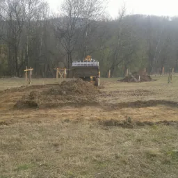 Żółta koparka przygotowuje teren pod budowę domu, widoczne wyznaczone fundamenty i drewniane słupki w trawie, w tle las.