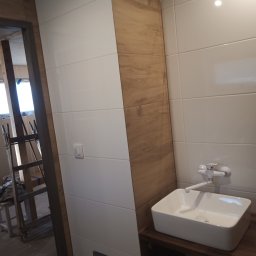 Dominik - Białe płytki ścienne w łazience z drewnopodobnymi akcentami, widoczna umywalka nablatowa i fragment rusztowania w tle.