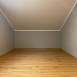 Poddasze po remoncie: świeżo pomalowane szare ściany, jasna drewniana podłoga, białe listwy przypodłogowe i sufitowe, widoczne gniazdka elektryczne i włącznik światła.