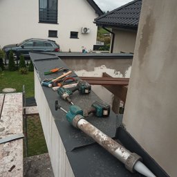 Dominik - Przybory dekarskie i pistolet do uszczelniania na czarnej obróbce blacharskiej, w tle budynek z białą elewacją i grafitową dachówką.
