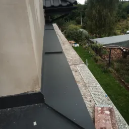 Fragment elewacji budynku z widocznymi obróbkami blacharskimi i fragmentem rusztowania na tle ogrodu z drzewami.