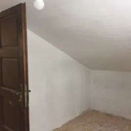 Poddasze po świeżym pomalowaniu na biało, widoczna stara drewniana stolarka drzwiowa i surowe wykończenie podłogi.