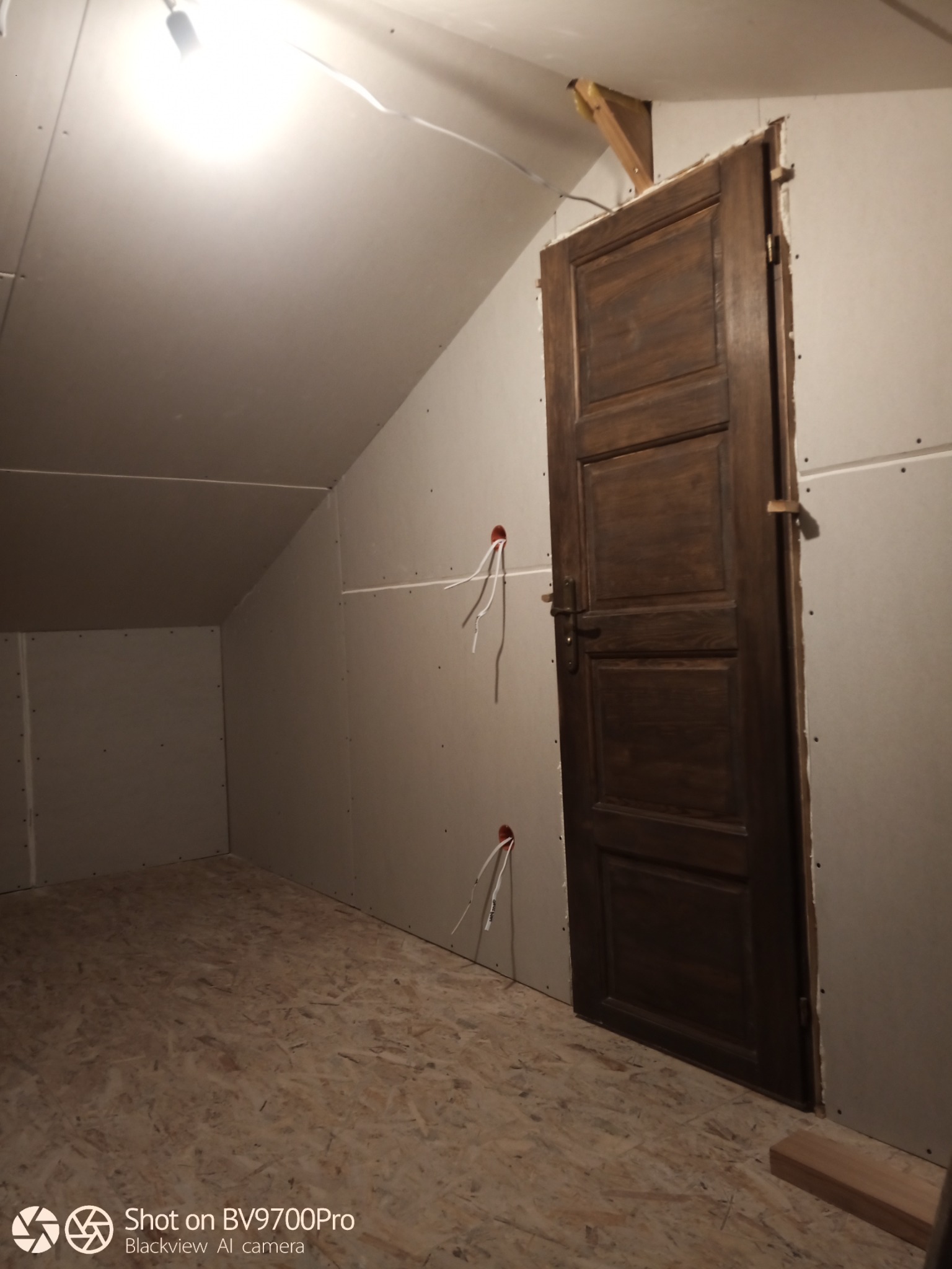 Poddasze w trakcie wykończenia: ściany obłożone płytami gipsowo-kartonowymi, widoczne przewody elektryczne, drewniane drzwi w ciemnym odcieniu brązu, podłoga OSB.