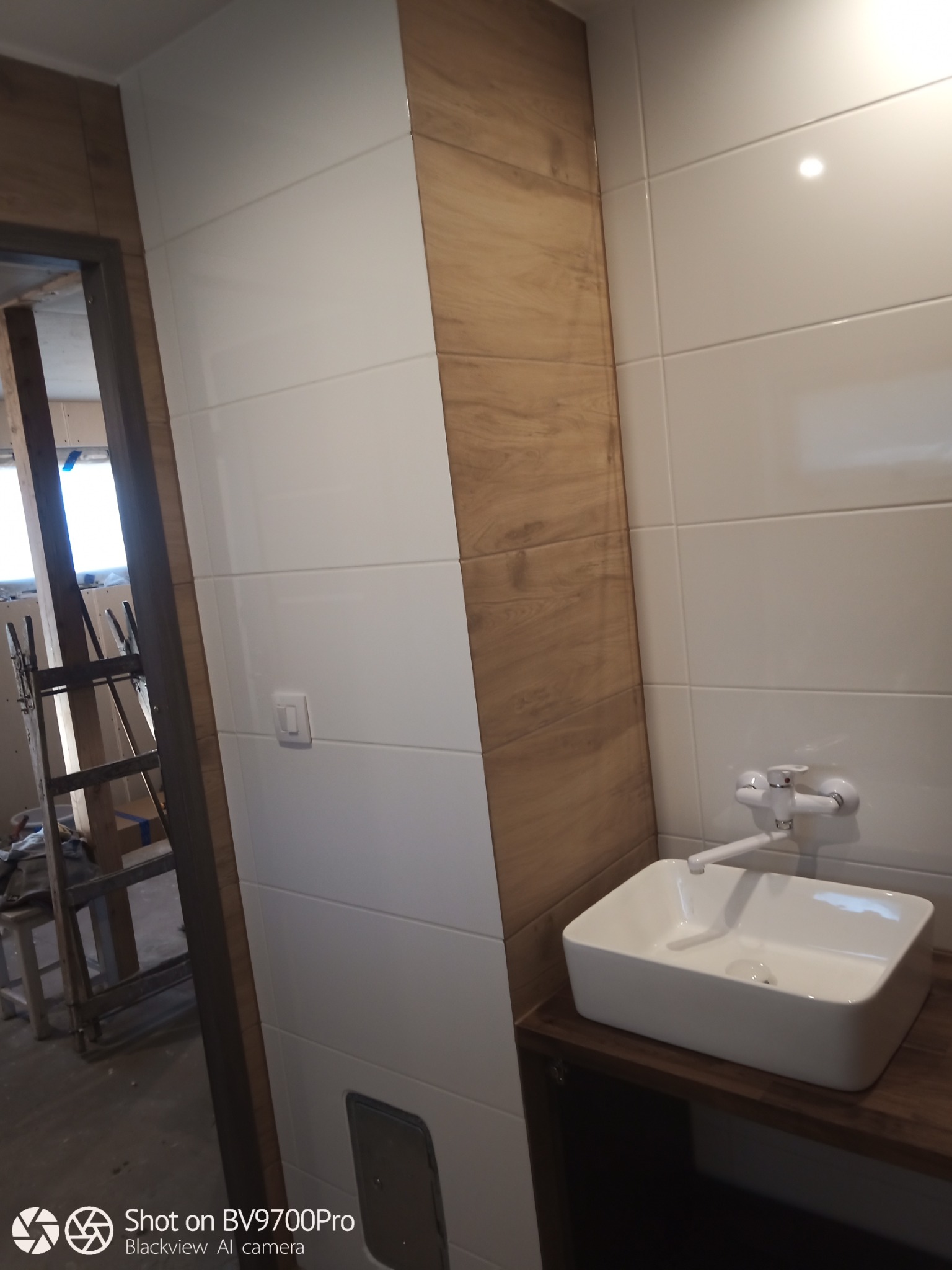 Białe płytki ścienne w łazience z drewnopodobnymi akcentami, widoczna umywalka nablatowa i fragment rusztowania w tle.