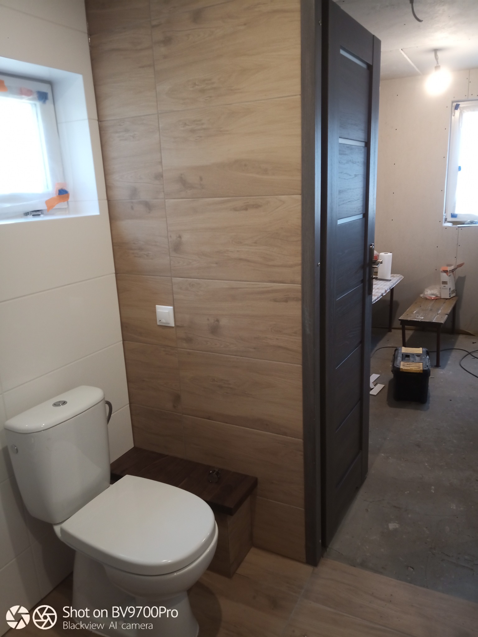 Nowoczesna toaleta z drewnopodobnymi płytkami na ścianie i ciemnymi drzwiami wewnętrznymi, widok na surowy stan wykończenia pomieszczenia.