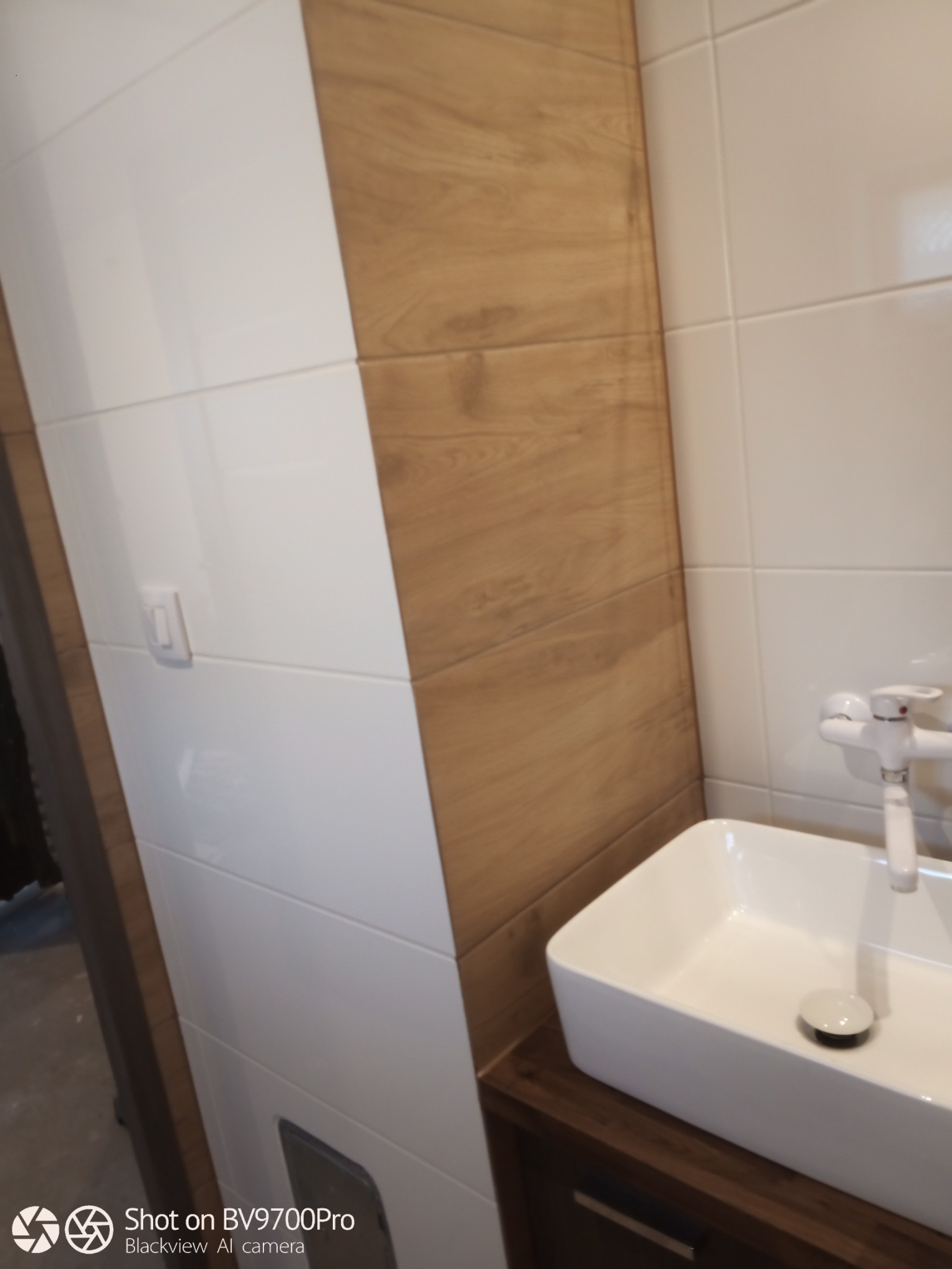 Nowoczesna łazienka z biało-drewnianymi płytkami na ścianach i minimalistyczną, kwadratową umywalką nablatową z białą baterią.