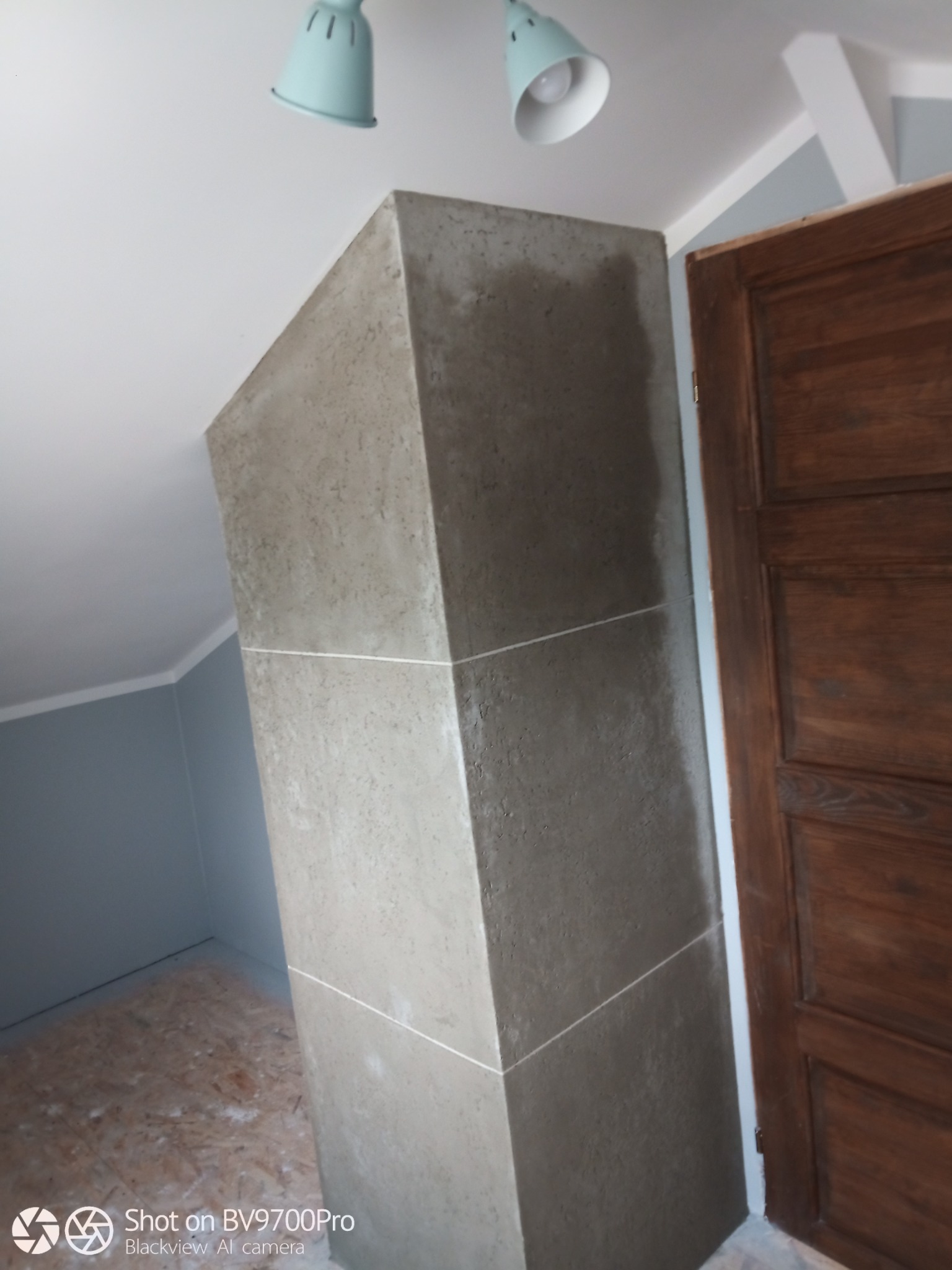 Wnętrze poddasza z kolumną wykończoną szarą fakturą, widoczna drewniana stolarka drzwiowa i lampy sufitowe w stylu retro.