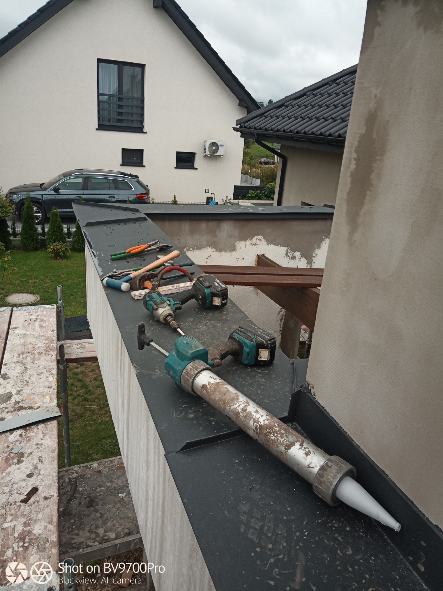 Przybory dekarskie i pistolet do uszczelniania na czarnej obróbce blacharskiej, w tle budynek z białą elewacją i grafitową dachówką.