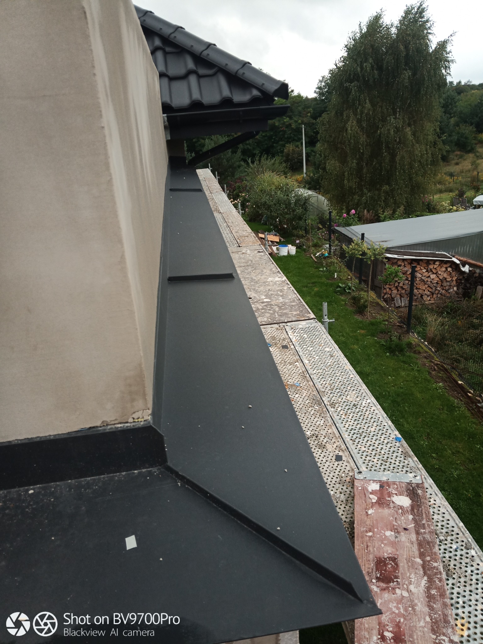 Fragment elewacji budynku z widocznymi obróbkami blacharskimi i fragmentem rusztowania na tle ogrodu z drzewami.