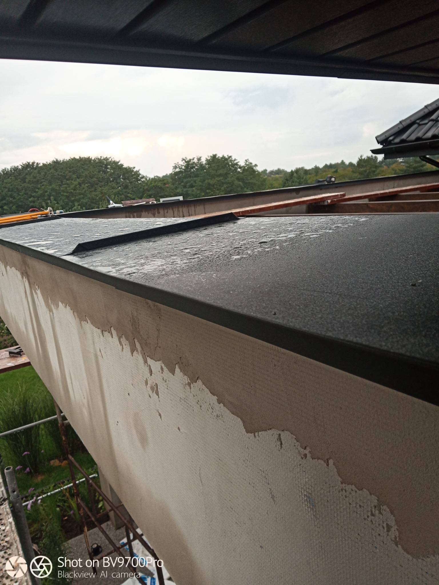 Fragment elewacji budynku w trakcie prac, widoczne mokre obróbki blacharskie na dachu i przygotowana powierzchnia ściany do dalszych prac wykończeniowych, z widokiem na zewnątrz i fragmentem...