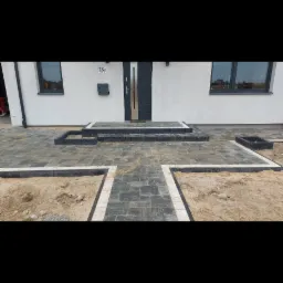 Ułożona ciemnoszara kostka brukowa przed nowoczesnym domem z białymi ścianami i ciemnymi oknami, widoczne przygotowane podłoże z piaskiem do dalszych prac.