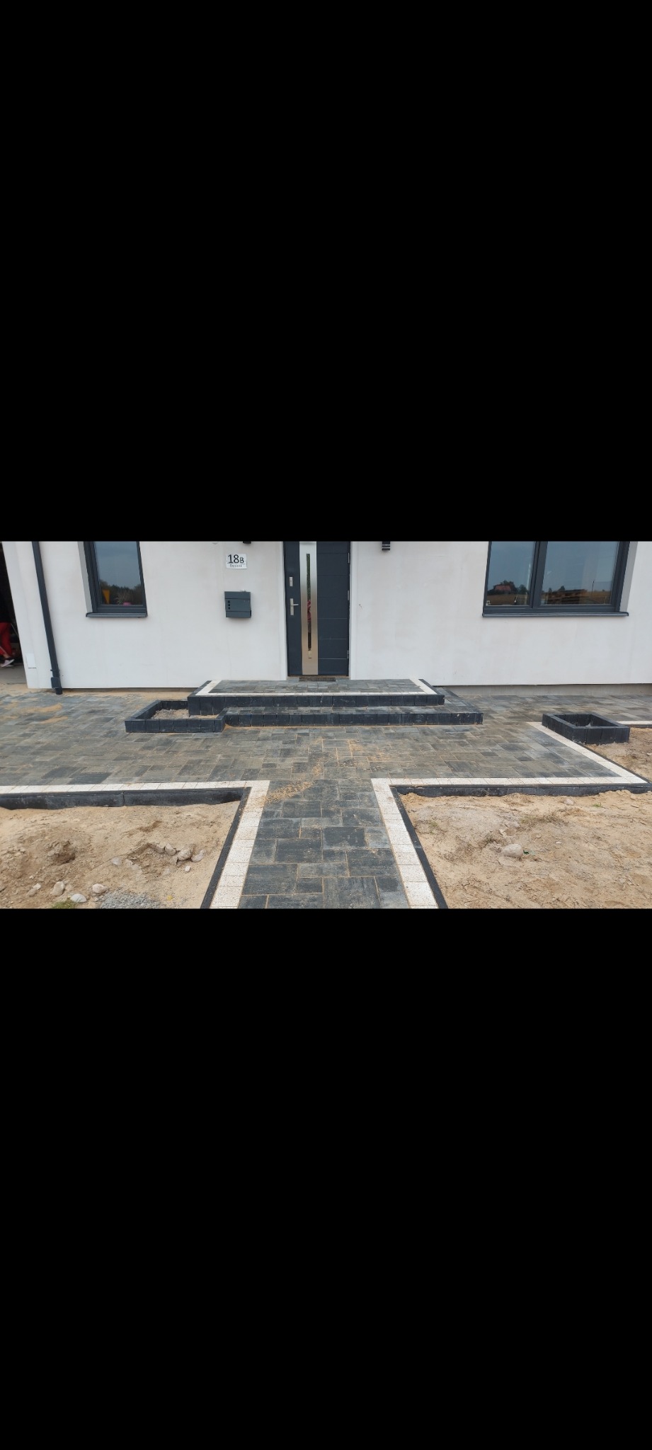 Ułożona ciemnoszara kostka brukowa przed nowoczesnym domem z białymi ścianami i ciemnymi oknami, widoczne przygotowane podłoże z piaskiem do dalszych prac.