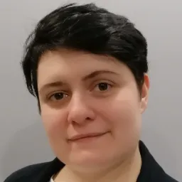 Portret kobiety z krótkimi czarnymi włosami, ubranej w białą koszulkę i czarną marynarkę, na tle szarej ściany.