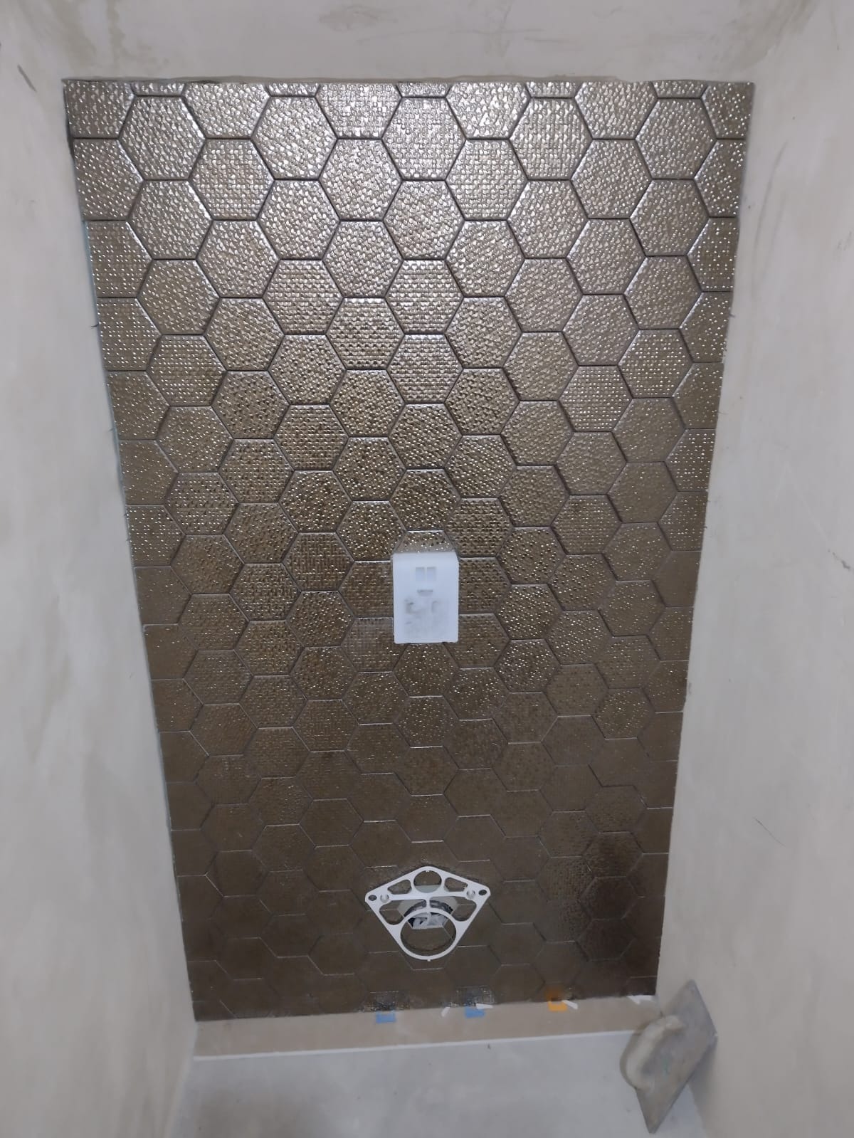 Ściana w łazience wyłożona brązowymi, heksagonalnymi płytkami z wyraźną teksturą, widoczny stelaż podtynkowy WC i element instalacji, ściany w stanie surowym.