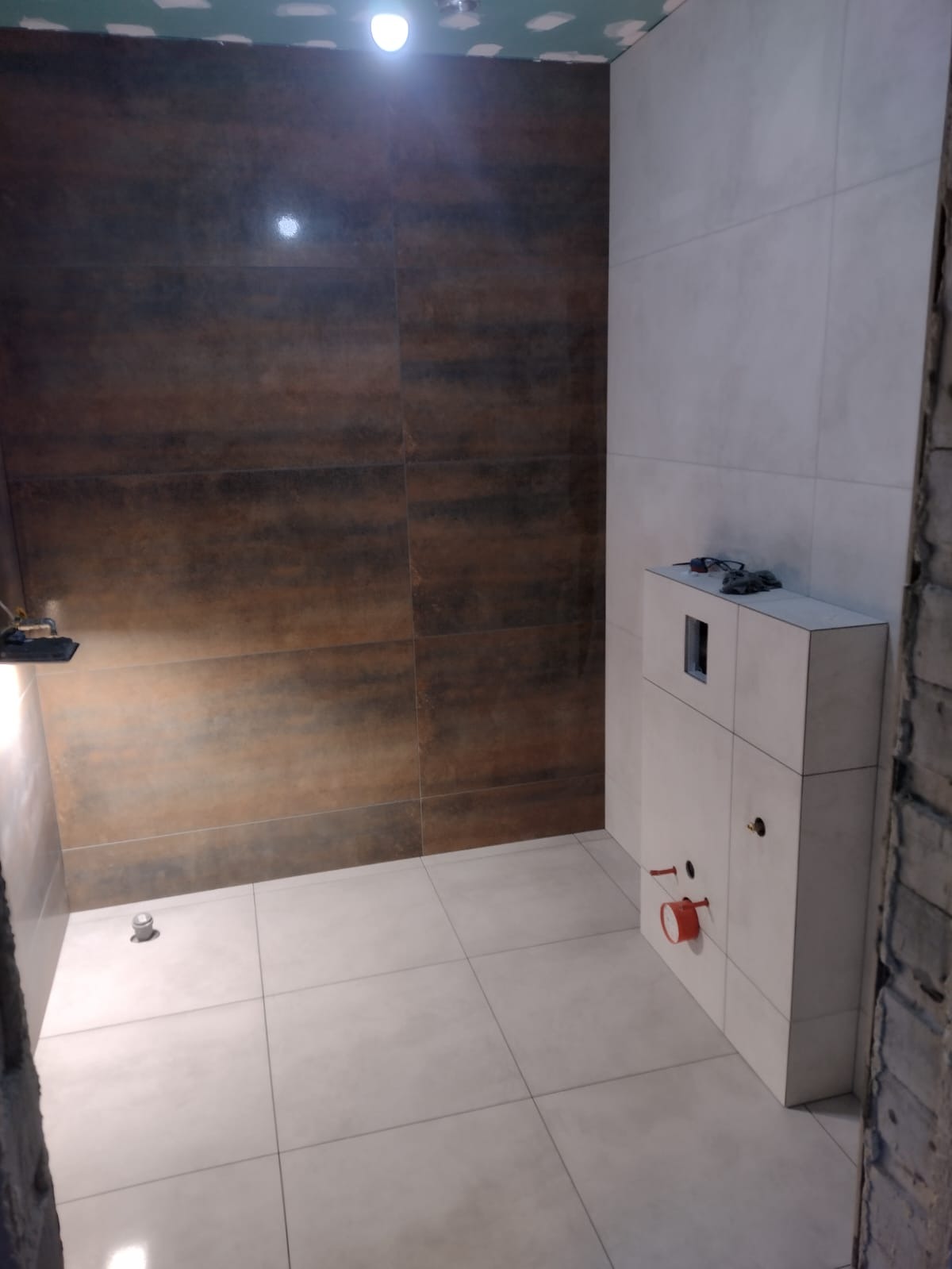 Łazienka w trakcie wykańczania, widoczne brązowe płytki imitujące drewno na ścianie, białe płytki na podłodze, zabudowa podtynkowego WC z akcesoriami hydraulicznymi, sufit częściowo wykończony...