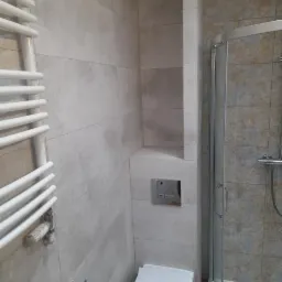 Wykończona łazienka z szarymi płytkami imitującymi kamień, zabudowaną wnęką w ścianie, grzejnikiem drabinkowym i kabiną prysznicową z chromowanymi profilami.