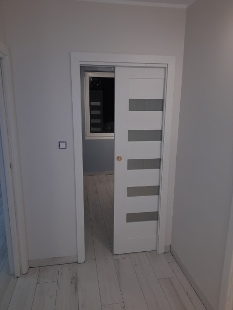 Białe przesuwne drzwi z poziomymi, szarymi paskami w otworze między pokojami, widoczne jasne panele podłogowe i fragment okna w tle.