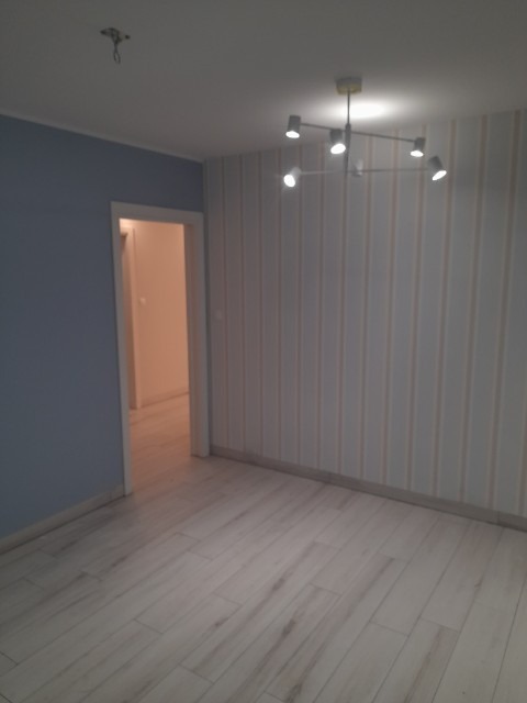 Pusty pokój po remoncie z panelami podłogowymi imitującymi drewno, niebieskimi ścianami, tapetą w pionowe paski i nowoczesną lampą sufitową z pięcioma reflektorami.