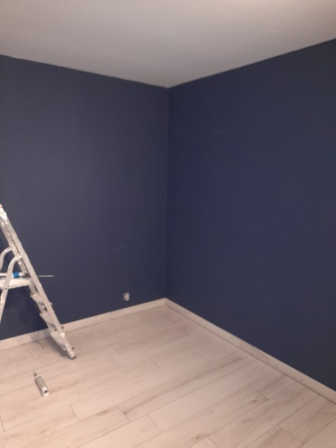Świeżo pomalowane na granatowo ściany w rogu pokoju z jasną podłogą, widoczna drabina malarska i puszka po farbie przy ścianie.