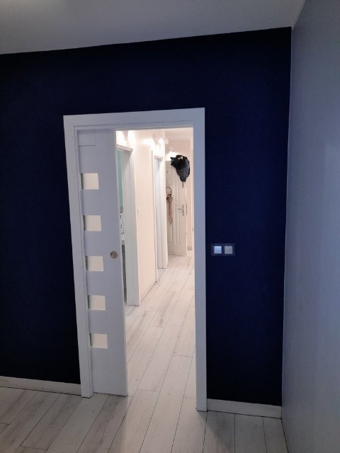 Wnętrze mieszkania po odświeżeniu: ciemnogranatowa ściana kontrastuje z białymi drzwiami z przeszkleniami i jasną podłogą z paneli. Widoczny fragment korytarza z białymi ścianami i drzwiami w głębi.