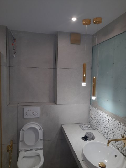 Nowoczesna łazienka z szarymi płytkami imitującymi beton, podwieszana toaleta z otwartą klapą, umywalka nablatowa z baterią w kolorze złotym i dekoracyjnym oświetleniem wiszącym.