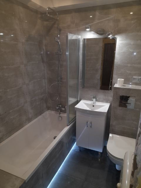 Wykończona łazienka z szarymi płytkami imitującymi kamień, podświetlana listwa LED pod wanną, biała szafka pod umywalką i lustro z oświetleniem punktowym.