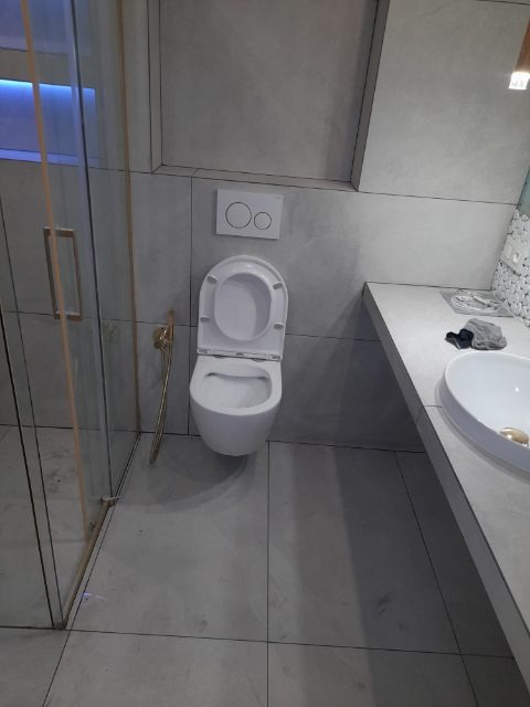 Nowoczesna toaleta podwieszana z otwartą deską, szare płytki podłogowe i ścienne, złoty uchwyt na szczotkę, minimalistyczna umywalka nablatowa i szklana kabina prysznicowa z niebieskim...