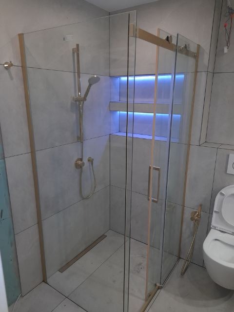 Nowoczesna kabina prysznicowa z przesuwnymi drzwiami, wykończona złotymi elementami, z wbudowanymi półkami podświetlonymi niebieskim światłem LED, widoczna toaleta w tle.