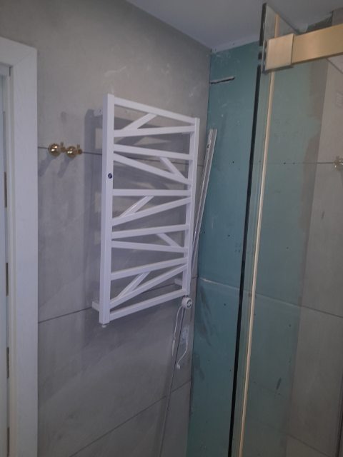 Biały, dekoracyjny grzejnik łazienkowy na szarej, wykończonej płytkami ścianie, obok kabiny prysznicowej w trakcie montażu.