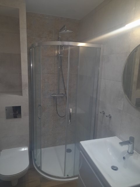 Wykończona łazienka z kabiną prysznicową o zaokrąglonym kształcie, białą umywalką z szafką i wiszącą toaletą w odcieniach szarości, widoczne chromowane baterie i okrągłe lustro.