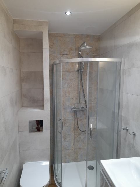 Wykończona łazienka z kabiną prysznicową z zaokrąglonymi szybami i deszczownicą, obok toaleta podtynkowa z chromowanym przyciskiem spłukującym, płytki w odcieniach beżu i szarości.