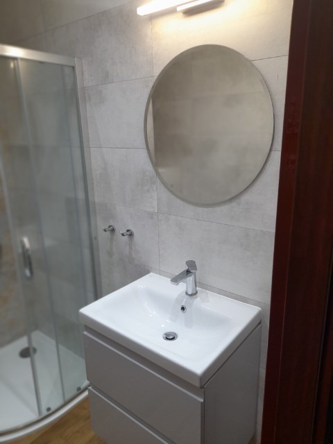 Nowoczesna łazienka z umywalką nablatową, okrągłym lustrem i kabiną prysznicową w tle, wykończona płytkami w odcieniach szarości.