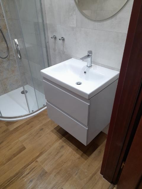 Nowoczesna łazienka po renowacji z drewnopodobną podłogą, białą umywalką z szafką, chromowaną baterią i kabiną prysznicową w tle.