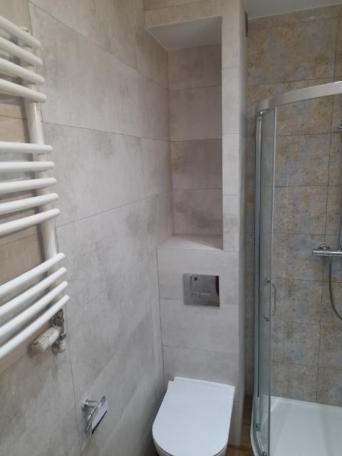 Wykończona łazienka z toaletą, grzejnikiem drabinkowym i kabiną prysznicową, wyłożona płytkami w odcieniach szarości i beżu z wnęką w ścianie.