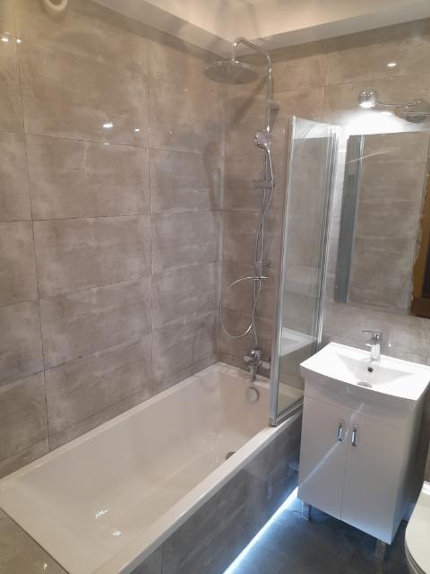 Wykończona łazienka z wanną, kabiną prysznicową typu walk-in i białą szafką pod umywalką, wykończona płytkami imitującymi marmur w odcieniach szarości.