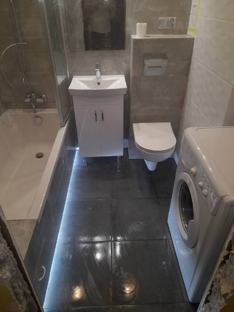 Wykończona łazienka z szarymi płytkami podłogowymi z podświetleniem LED, białą armaturą (umywalka z szafką, toaleta) i pralką.