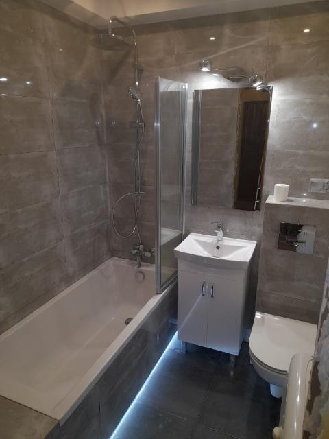 Wykończona łazienka z szarymi płytkami imitującymi kamień, oświetleniem LED pod wanną, białą szafką pod umywalką i kabiną prysznicową ze szklanymi drzwiami.