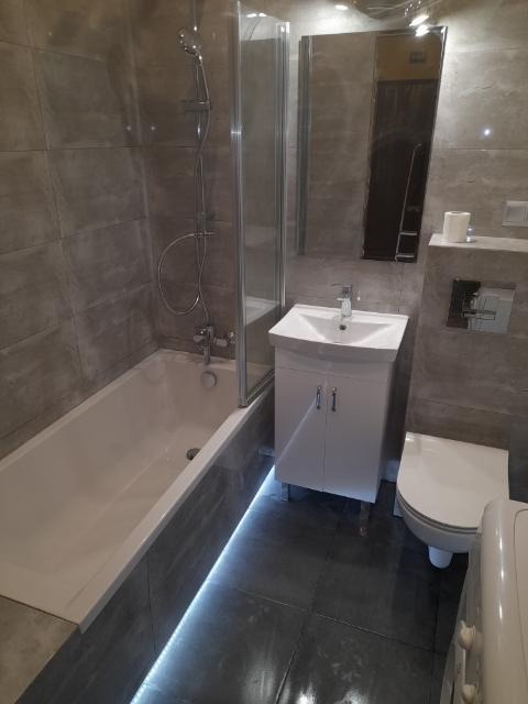 Wykończona łazienka z szarymi płytkami, białą wanną z parawanem, umywalką z szafką i toaletą, oświetlona listwą LED pod wanną.