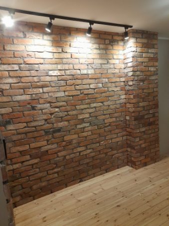 Ściana z czerwonej cegły z oświetleniem punktowym na suficie i jasną drewnianą podłogą, widok na róg pomieszczenia.