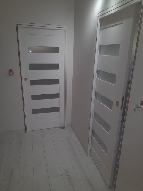 Dwa białe, nowoczesne drzwi wewnętrzne z poziomymi, matowymi przeszkleniami, zamontowane w jasnym pomieszczeniu z panelami podłogowymi o strukturze drewna.