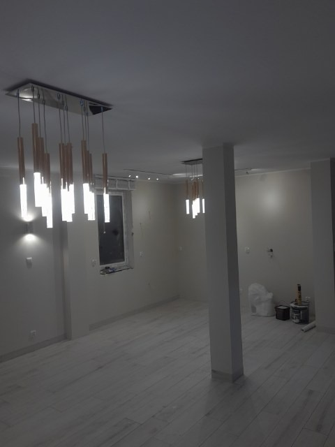 Wnętrze po remoncie w Wejherowie z nowoczesnym oświetleniem: podłużne lampy wiszące z drewnianymi elementami, jasne ściany i podłoga imitująca drewno, widoczne puszki z farbą w rogu pomieszczenia.