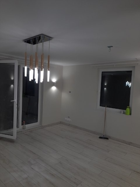 Wykończone wnętrze pokoju z drewnopodobną podłogą, nowoczesnym oświetleniem sufitowym z podłużnych lamp, otwartym balkonem i oknem z widokiem na zewnątrz. Na parapecie stoi zielona butelka, a przy...