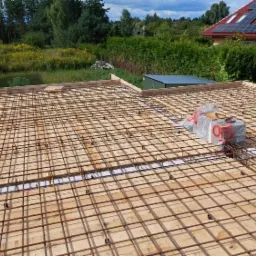 Zbrojenie płyty fundamentowej na deskowaniu, widoczne stalowe pręty tworzące siatkę, dystanse pod zbrojeniem, fragment budynku w tle.