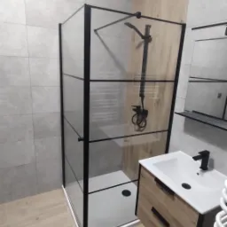 Nowoczesna łazienka z kabiną prysznicową typu walk-in z czarnymi profilami, umywalką nablatową z szafką w okleinie drewnopodobnej i czarną armaturą, wykończona płytkami w odcieniach szarości i drewna.