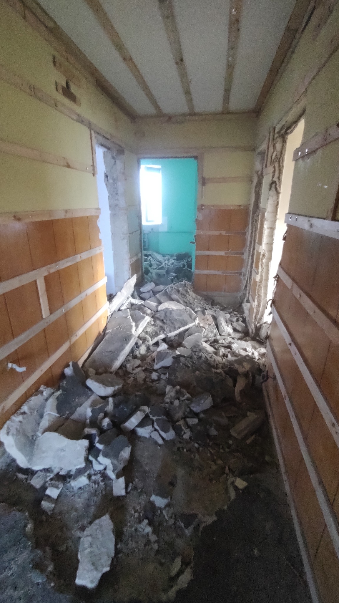 Korytarz w trakcie demontażu, widoczne fragmenty gruzu i odsłonięte deski na ścianach, w tle turkusowe drzwi.