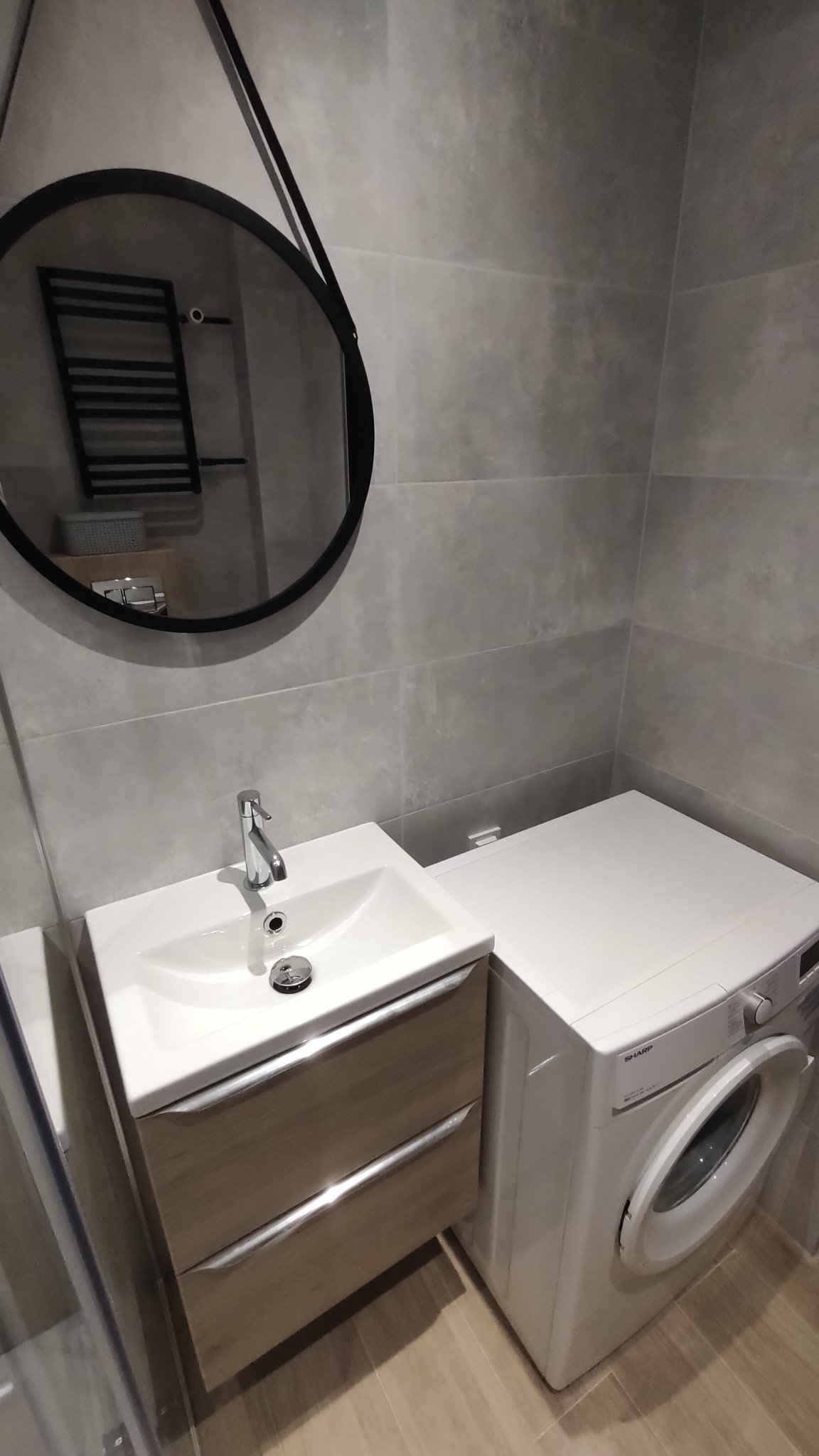 Nowoczesna łazienka w szarościach z umywalką na szafce z drewnianymi szufladami i pralką Sharp, widoczna okrągła rama lustra z odbiciem grzejnika.